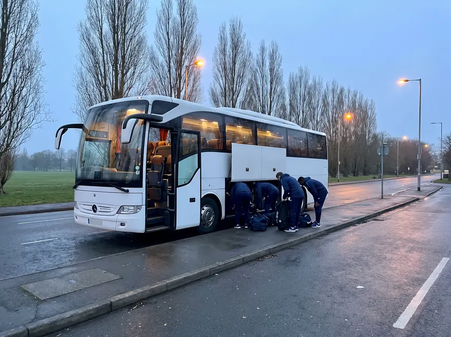 A white touring coach at the roadside beside open meadow and bare poplar trees at dawn, street lamps still lit, a sports team in navy tracksuits loading kit bags into the luggage bay